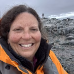A person smiles at the camera wearing an orange jacket and life vest, standing on rocky terrain under a cloudy sky. Another figure is visible in the distance on the rocks.